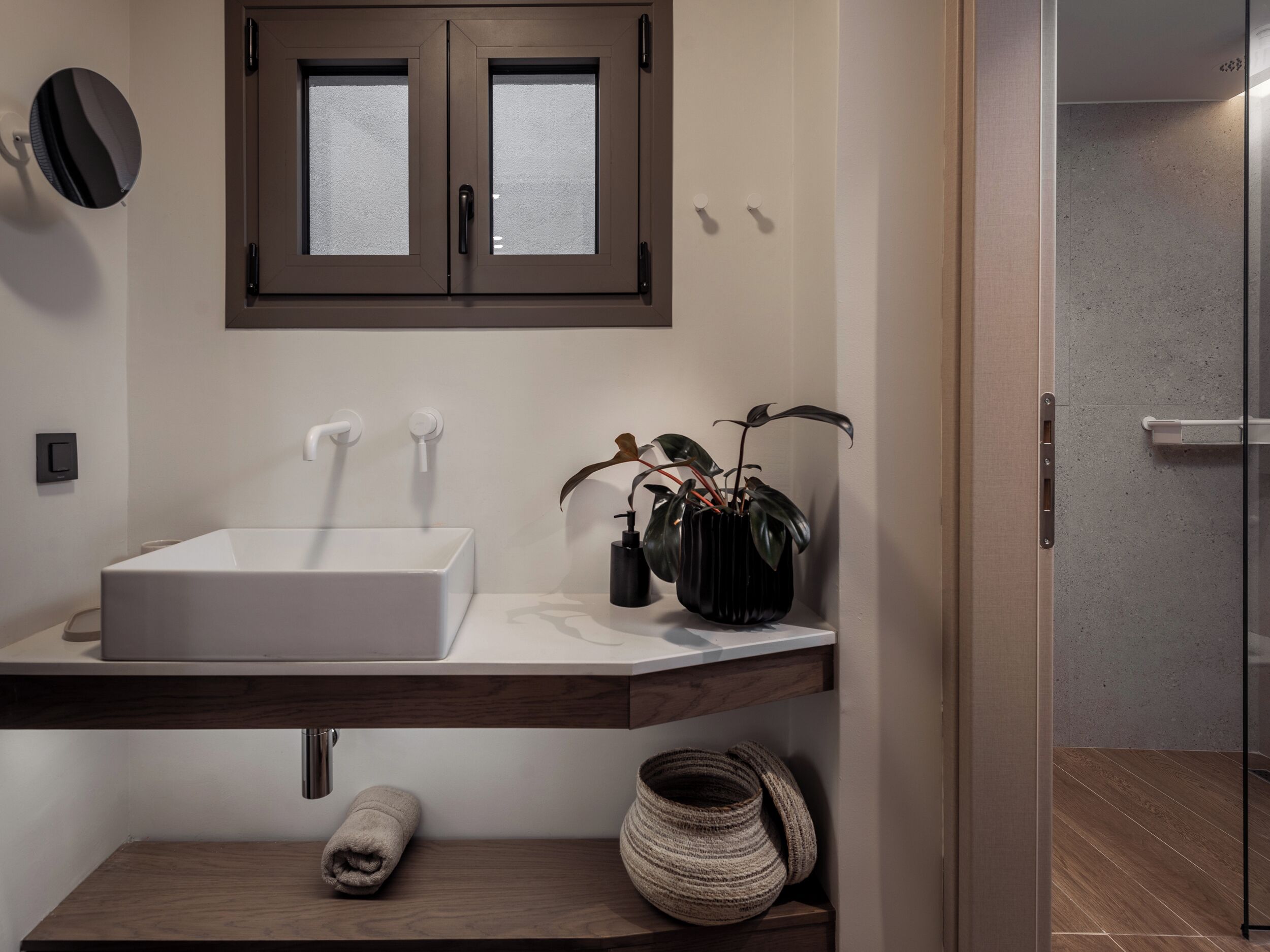 Un baño moderno con un lavamanos blanco, dos dispensadores de toallas, una planta en maceta y un estante de madera con una toalla enrollada y una cesta tejida.