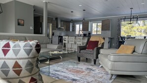 A spacious living area with a sofa, armchair, and coffee table, a kitchen in the background, and a large window providing natural light. - INDOOR POOL, HOT TUB & SAUNA!!  Welcome to Bluehaven in the Kawartha's!! (Buckhorn)