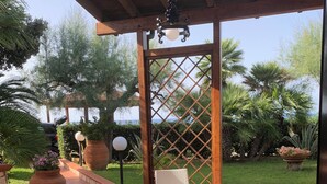 Ein überdachter Patio-Bereich mit Korbmöbeln, einem runden Glastisch und Blumentöpfen.