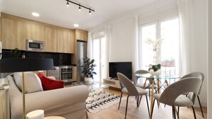 Un salon moderne avec un canapé, une table basse et un téléviseur. Il y a également un coin cuisine équipé d’un micro-ondes, d’un four et d’un réfrigérateur. La pièce est dotée de grandes fenêtres et offre une vue sur la ville.