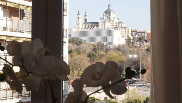 Une vue d’une ville avec une imposante cathédrale en arrière-plan, une rue bordée de voitures garées et un immeuble avec des balcons au premier plan.