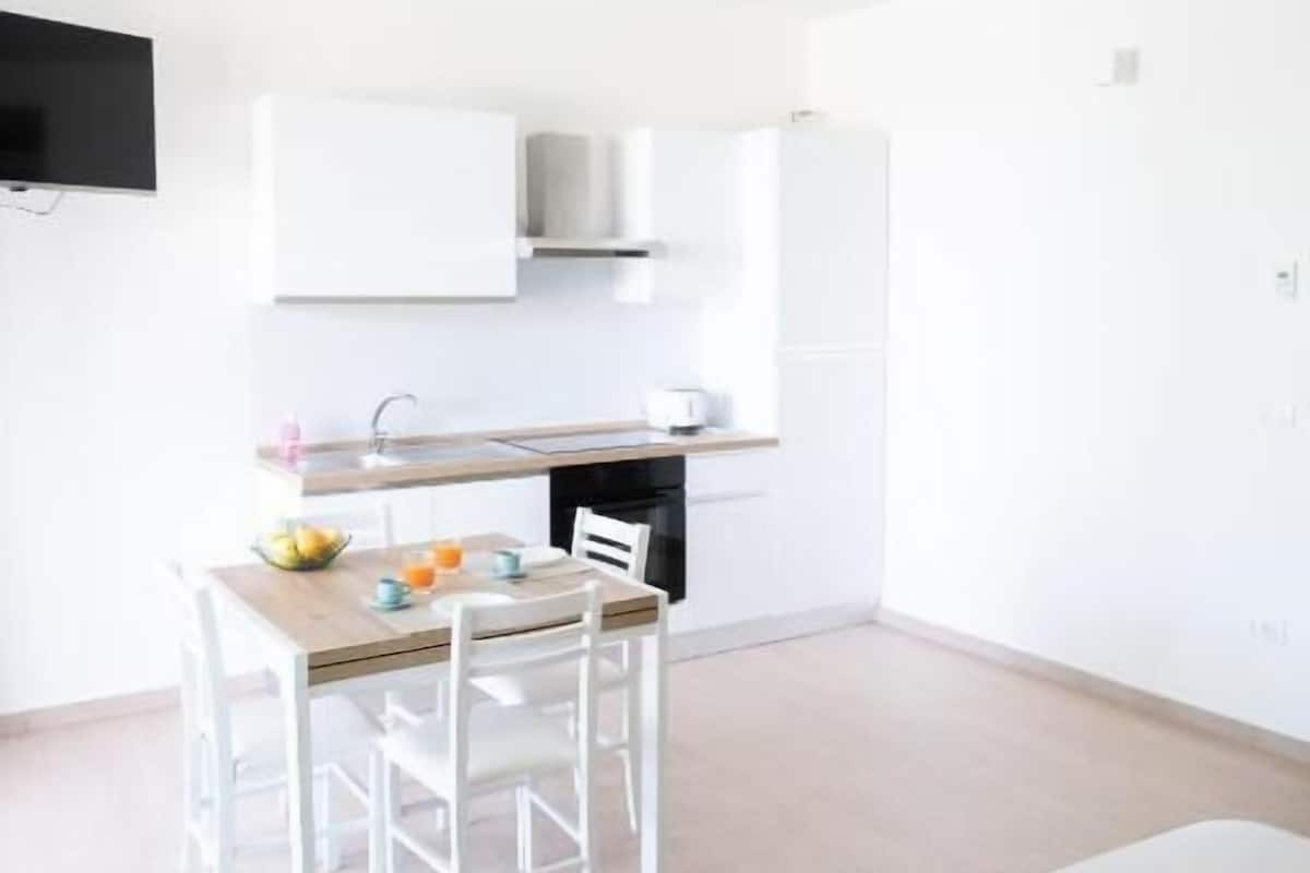 A modern, minimalist hotel room with a neatly made bed, a small dining table with chairs, a kitchenette, and a flat-screen TV mounted on the wall.