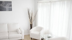 Ein minimalistisches Wohnzimmer mit einem weißen Sofa, einem Glastisch und zwei Sesseln. Zwischen Sofa und Sesseln steht eine Vase mit Zweigen.