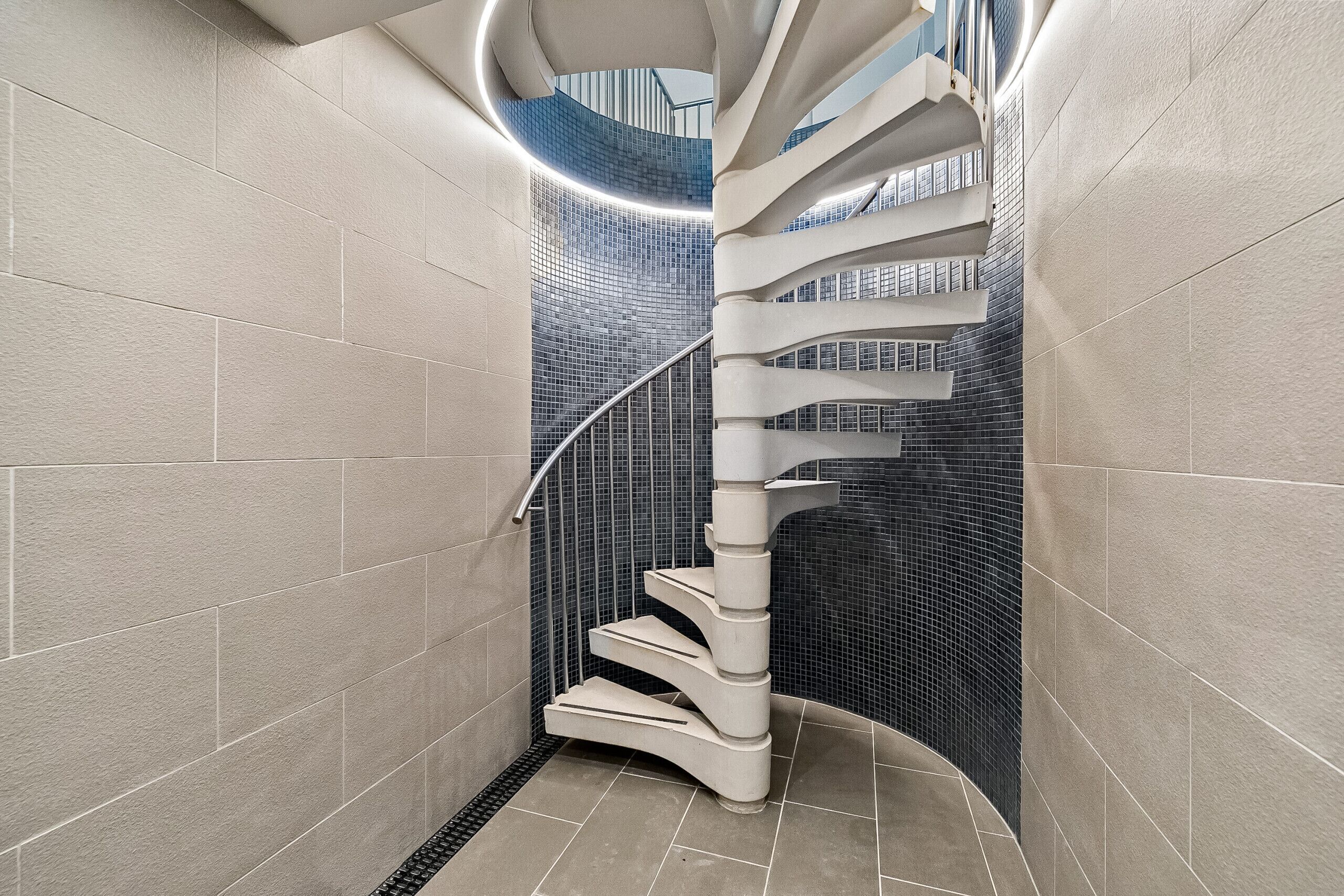 Un escalier en colimaçon au design moderne, entouré de murs aux couleurs claires et surmonté d’un sol carrelé foncé.