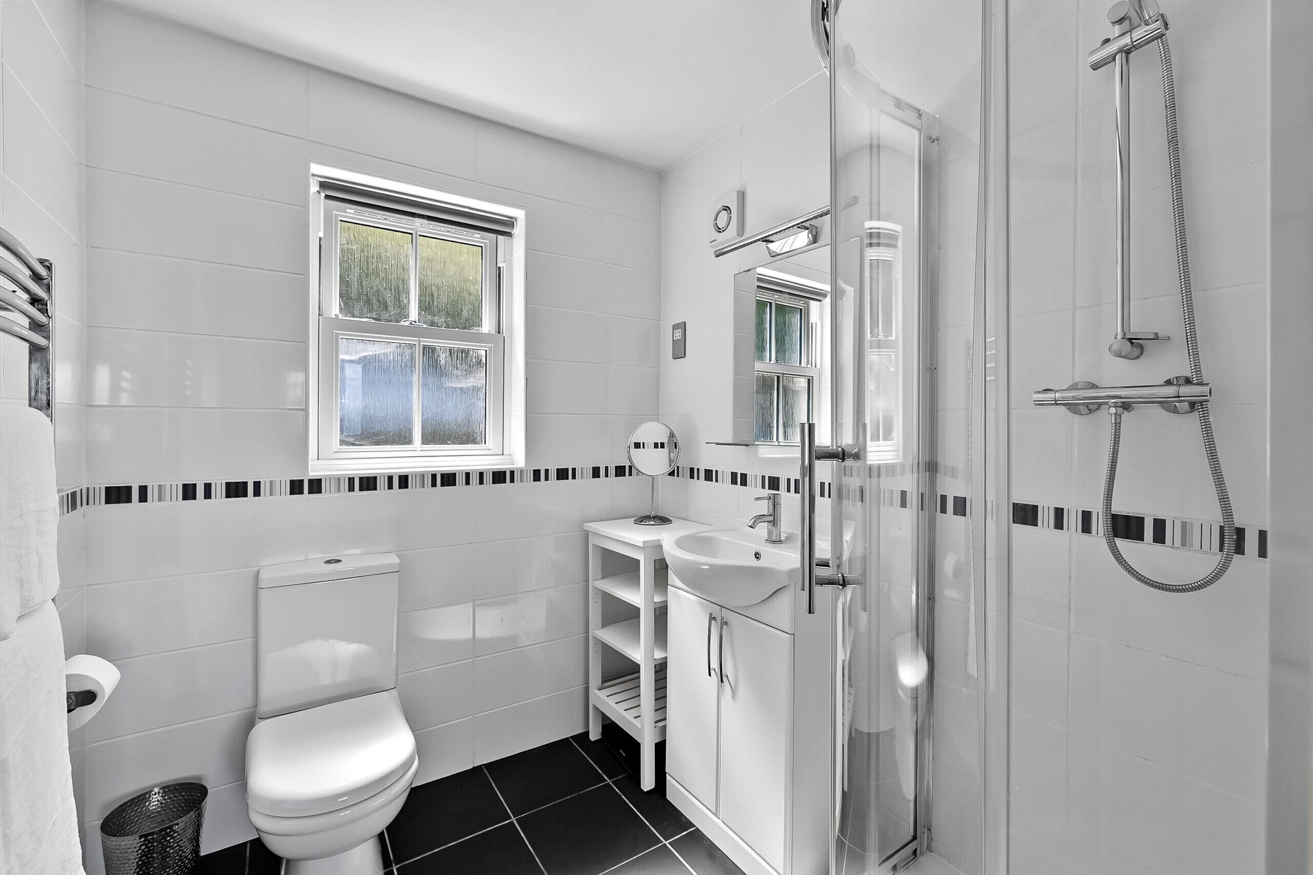 A modern bathroom with a toilet, sink, and shower. There is a window above the toilet and a mirror above the sink.