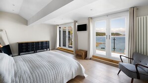 Een slaapkamer met een groot bed, een aan de muur bevestigde televisie en uitzicht op zee door grote ramen.