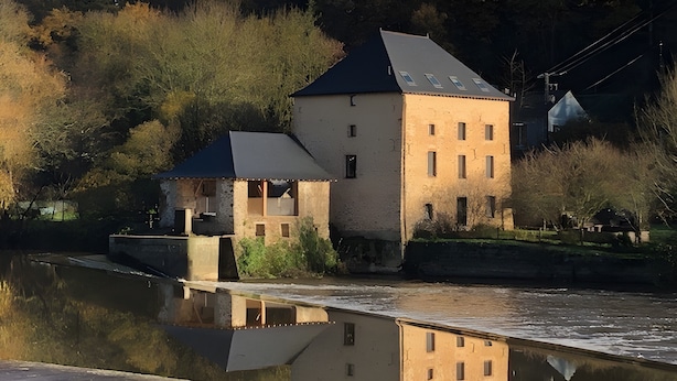 Un bâtiment situé au bord de la rivière, avec un petit quai, un pont et son reflet dans l’eau.