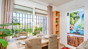 Uma área de refeições com uma mesa posta para dois, uma planta em vaso e vista para uma varanda com guarda-corpo.