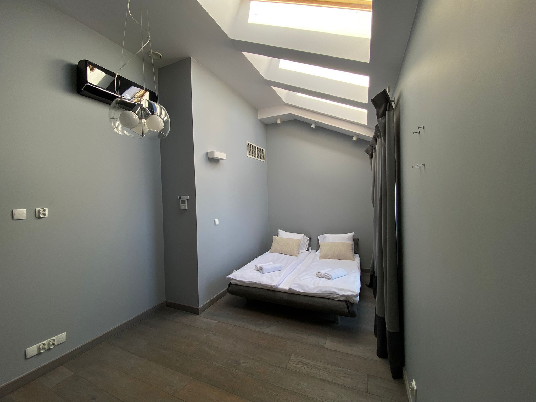 Una habitació estreta amb una finestra al sostre, un llit i un mirall a la paret.