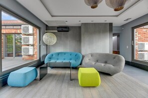 A modern living room with a blue sofa, a grey sofa, and a yellow ottoman. There are air conditioning units on the windows. - Tobiasza by Browar Hevelius Apartments (Gdańsk)