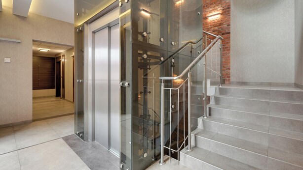 A modern interior with a glass elevator, a staircase with metal railings, and a brick wall. - Tobiasza by Browar Hevelius Apartments (Gdańsk)