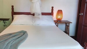 A bed with white linens, a wooden headboard, a bedside table with a lamp, and a door to the right. - Modest family home located in a quiet neighbourhood on Calibishie Ridge. (Calibishie)