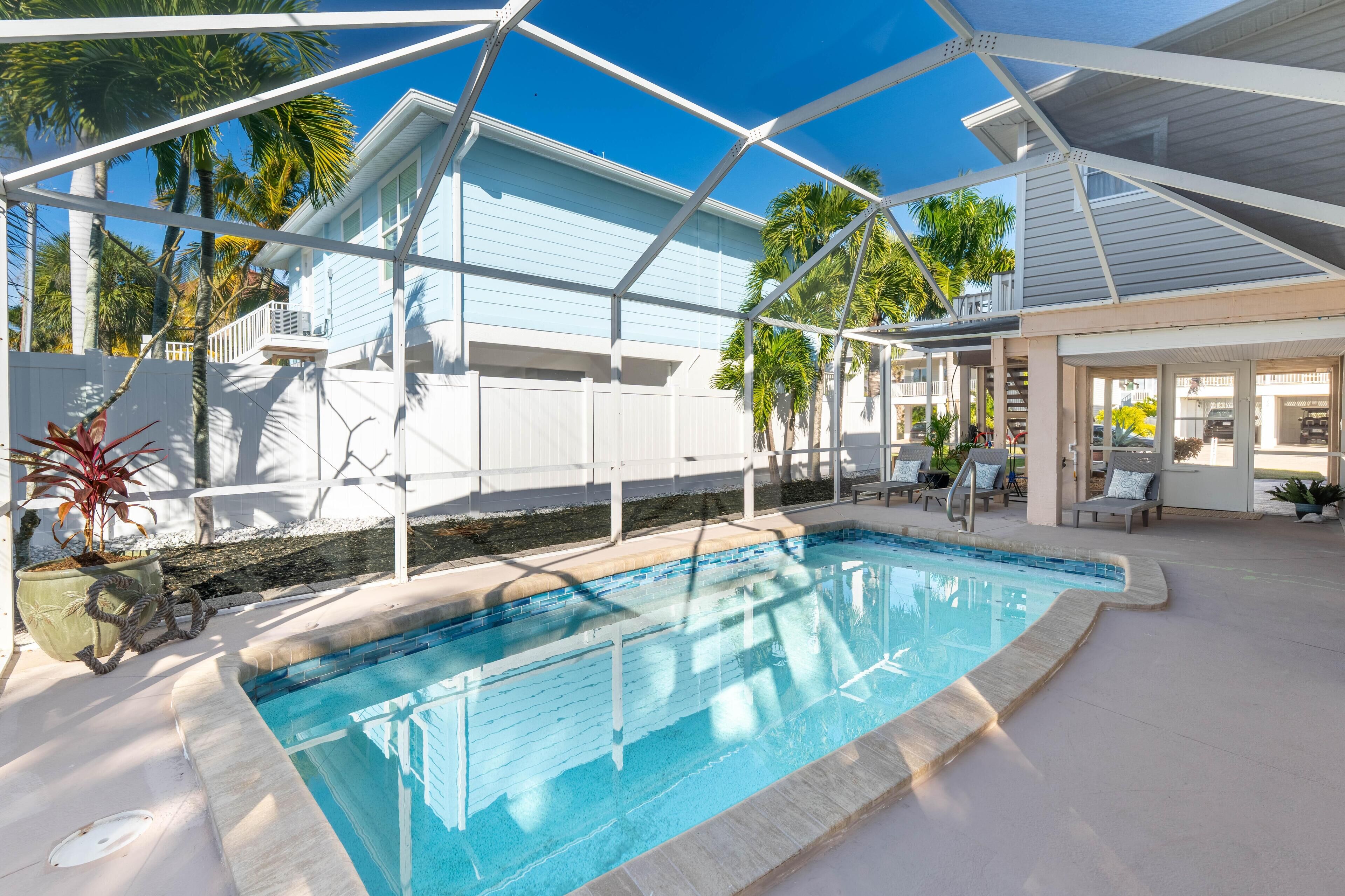 Un área de piscina con una piscina azul cristalina, sillas blancas junto a la piscina y un patio cubierto con vistas a una casa azul claro y palmeras.