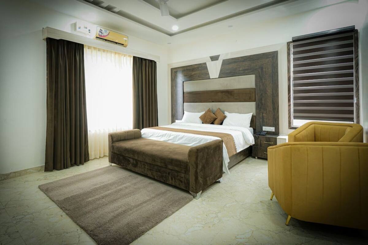 Una camera d'albergo con un letto, un divano, una sedia, una finestra con tende, un condizionatore d'aria e un termosifone.