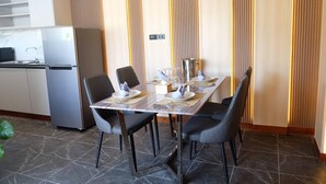 Un coin repas moderne avec une table rectangulaire dressée pour quatre personnes, entourée de chaises noires. On aperçoit un réfrigérateur et un four à micro-ondes à l’arrière-plan.