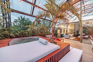 Un jardín interior moderno con una bañera de hidromasaje, asientos de madera y una zona de estar cubierta con paredes de vidrio.