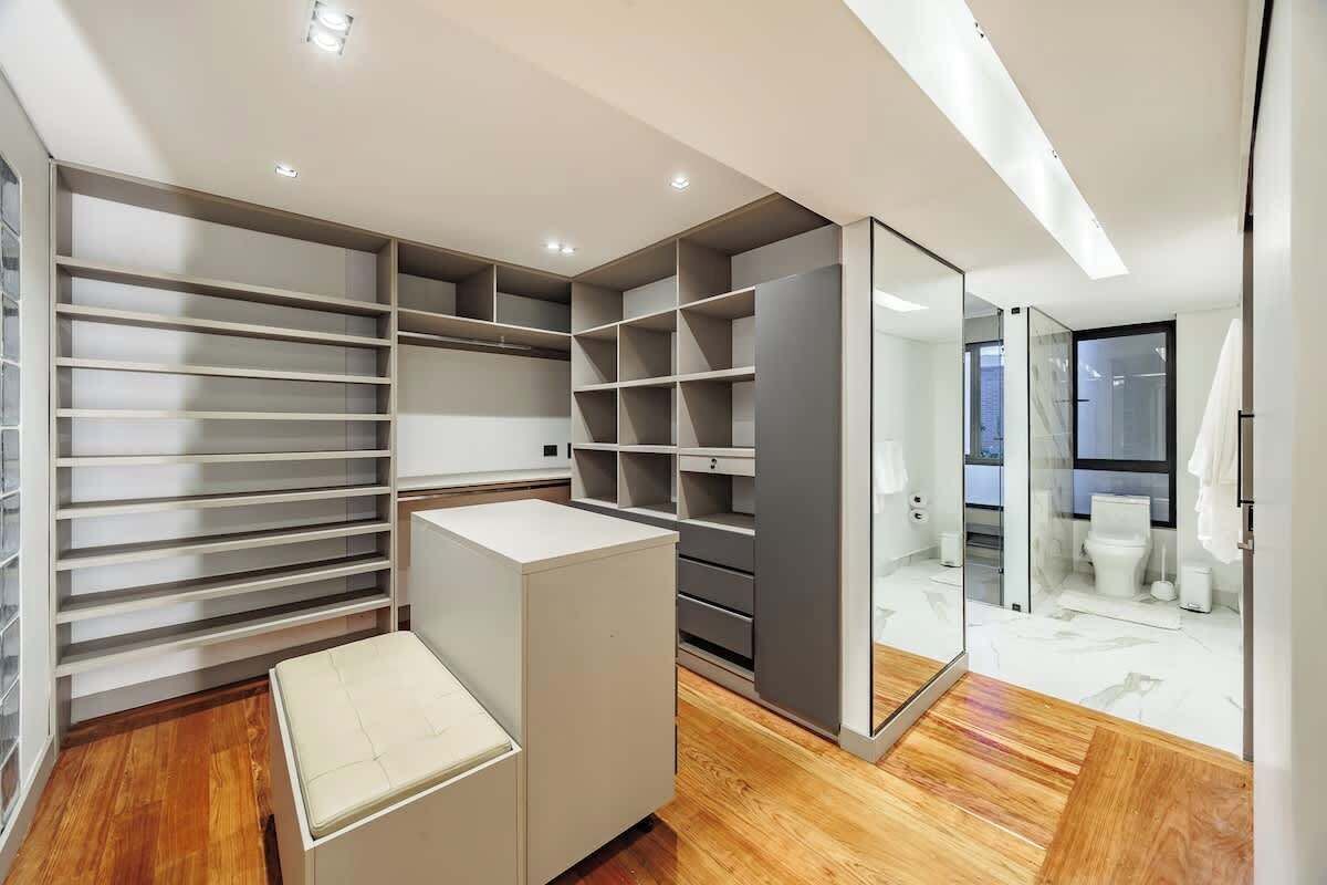 A modern closet with multiple shelves and drawers, a bench, and a glass partition leading to a bathroom.