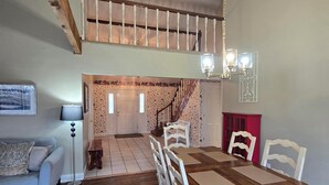 Un área de comedor con una mesa de madera y sillas blancas, una escalera que sube a un nivel superior y una alfombra estampada en el suelo.