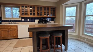 Una cocina con armarios de madera, un lavavajillas blanco, una barra con taburetes y ventanas amplias.