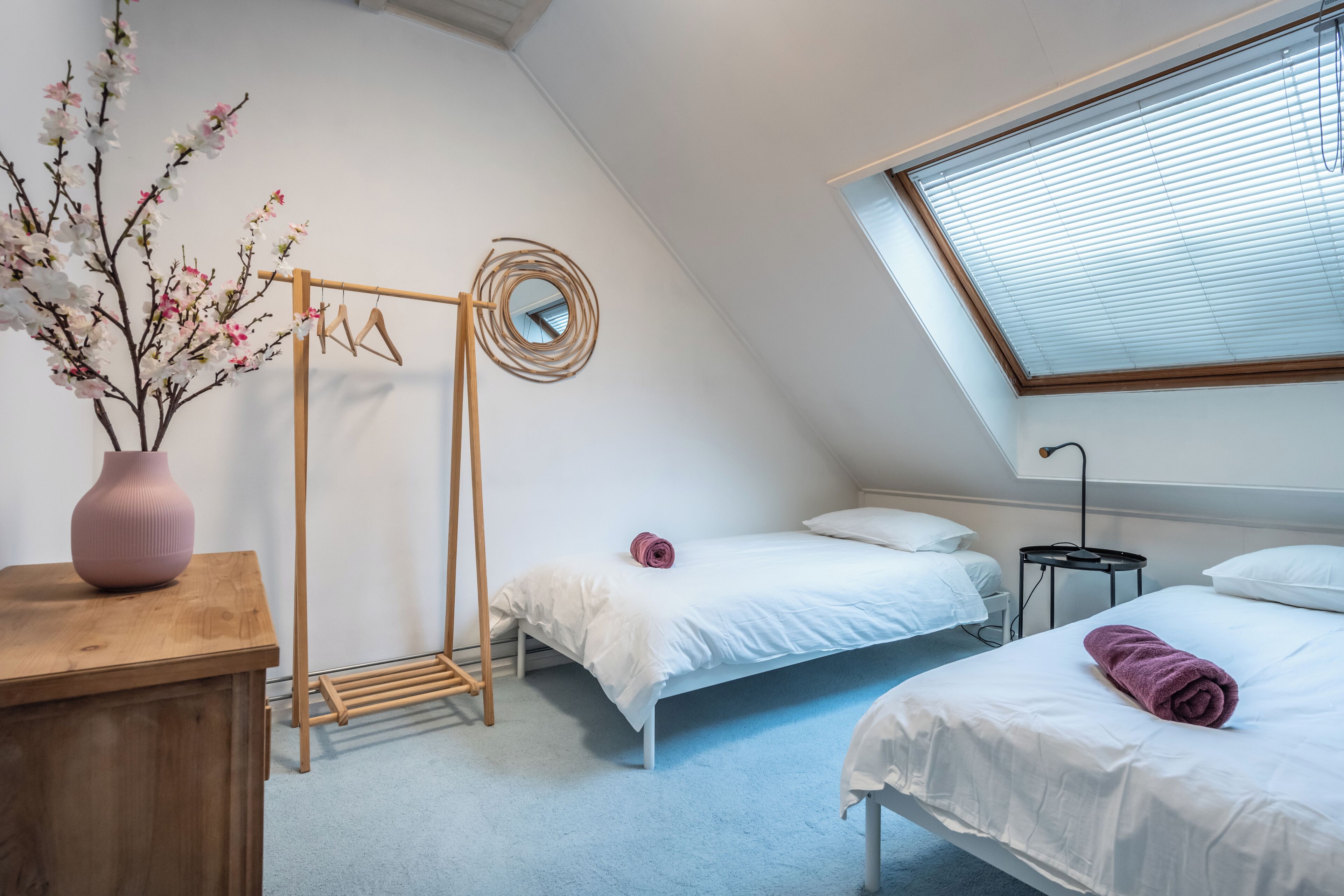 A bedroom with a sloped ceiling, a wooden dresser, a hanging clothes rack, two beds with white bedding, a small round mirror, and a window with blinds.
