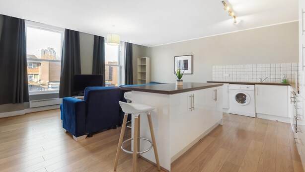 A modern kitchen with a built-in washing machine, a bar area with stools, and a large window with curtains. - Great 2-Bed Flat in Prime Location (Greater London)