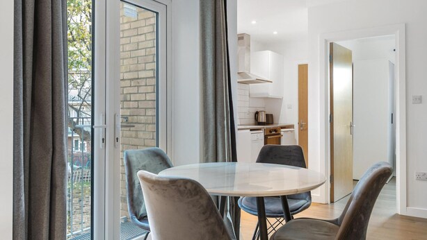 A modern apartment with a dining area featuring a round table and four chairs, a kitchen with white cabinets, and a sliding glass door leading to a balcony. - Modern Flat in Prime Location (Greater London)