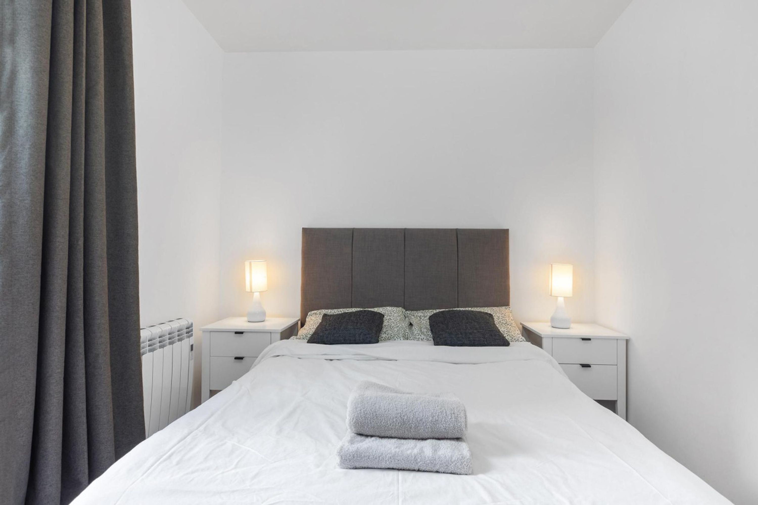 Ein ordentlich bezogenes Bett mit weißer Daunendecke und zwei Kissen, an beiden Seiten mit Nachttischen mit Lampen, in einem Zimmer mit schlichter weißer Wand und grauem Vorhang.