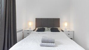 A neatly made bed with a white duvet and two pillows, flanked by two bedside tables with lamps, in a room with a plain white wall and a grey curtain. - Modern Flat in Prime Location (Greater London)