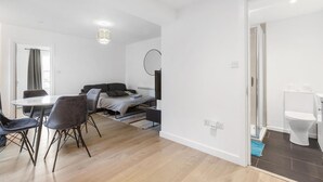 A modern, well-lit room with a dining table, chairs, a sofa, and a bed. A small circular mirror hangs on the wall. - Modern Flat in Prime Location (Greater London)