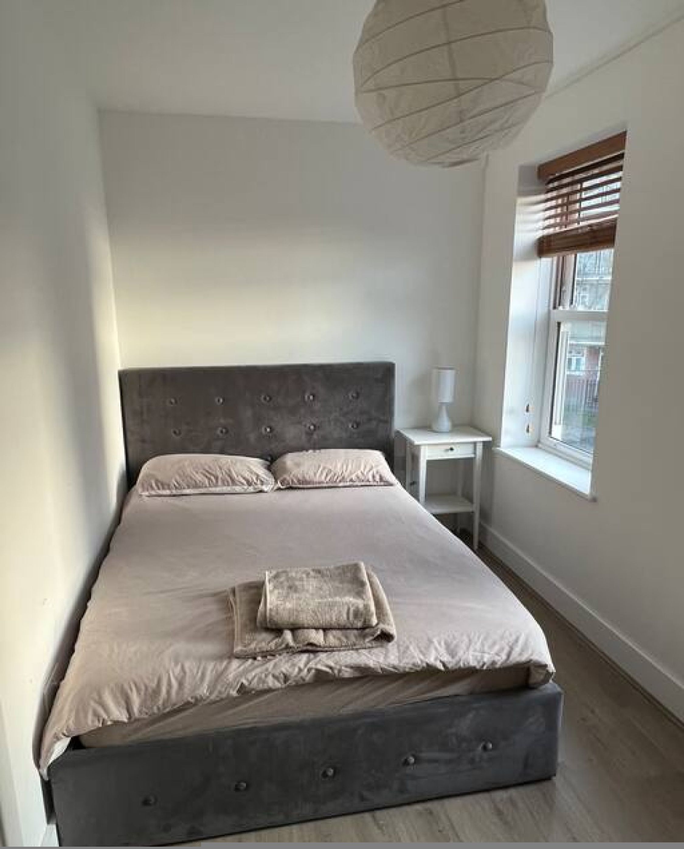A bedroom with a large bed, a bedside table, a lamp, and a window with blinds.