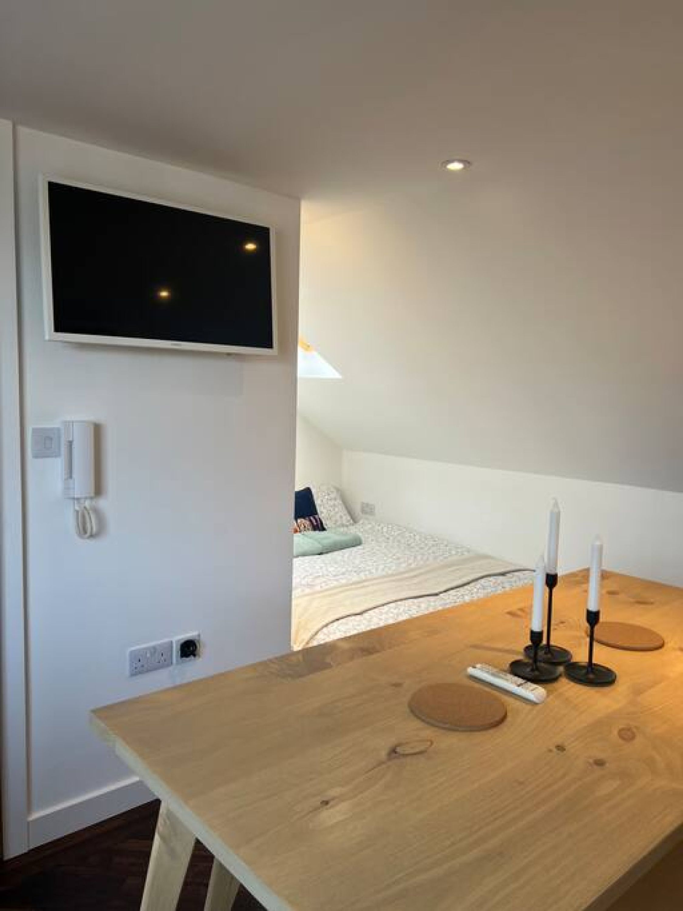 A room with a bed, a TV mounted on the wall, a wooden table with candles, and a small table lamp.