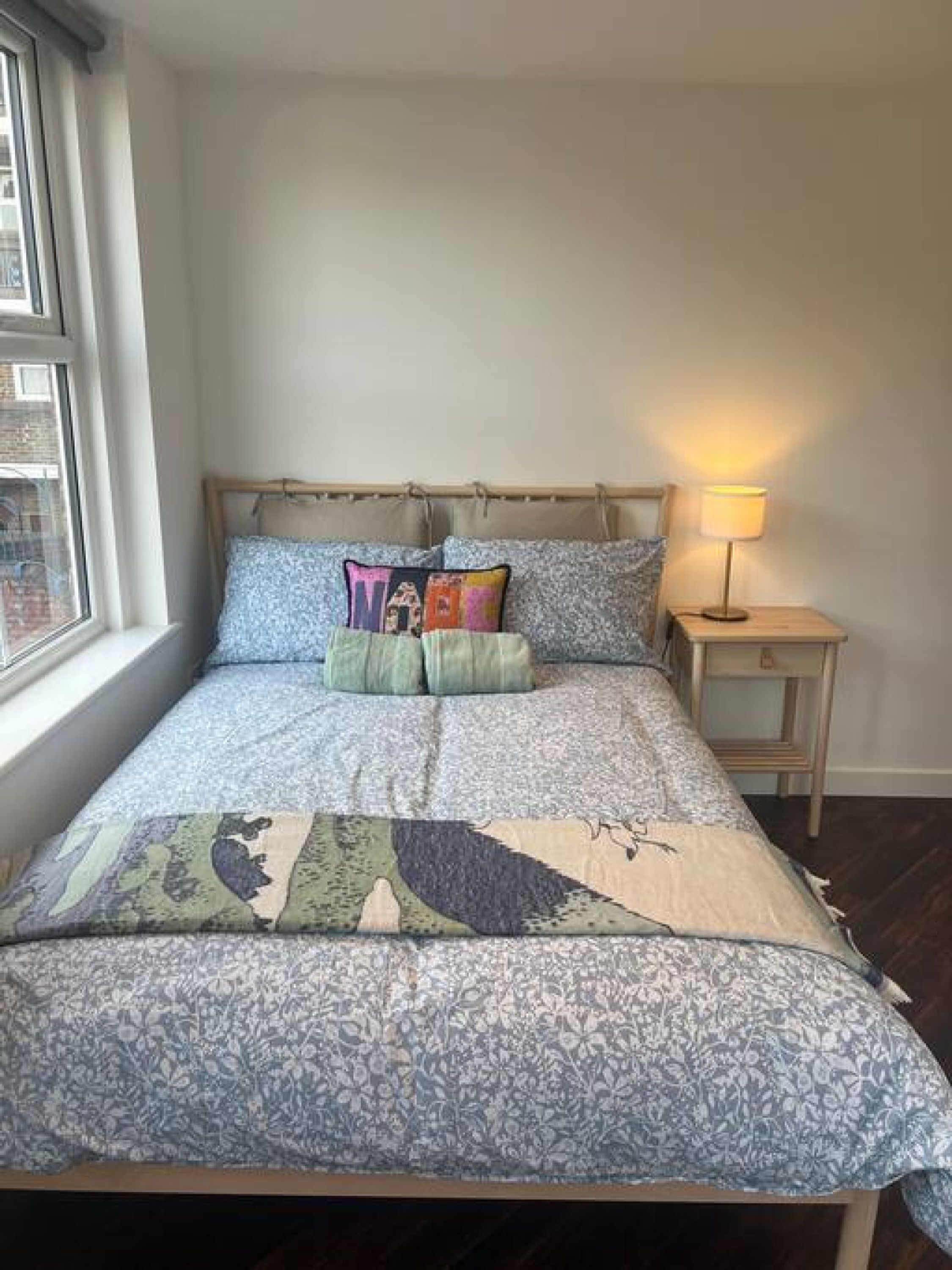 Un lit avec une couvre-lit à motifs, plusieurs oreillers, une lampe de chevet et une fenêtre avec des rideaux.