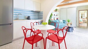 Eine moderne Küche mit weißen Schränken, einer gestreiften Spritzschutzwand und einem Edelstahl-Kühlschrank. Ein runder Tisch mit roten Stühlen und eine Schüssel mit grünen Äpfeln stehen bereit. Die Küche geht in einen Wohnbereich mit einem blauen Sofa und einem Couchtisch über.
