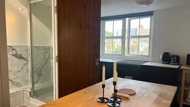 A modern kitchen with a wooden table, two candles, a coffee maker, and a view of the city through a large window. - Spacious Ensuite Room in Clapham (Greater London)