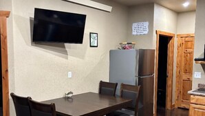 A dining area with a table, chairs, a television mounted on the wall, and a refrigerator. - Hungry Mosquito lodge 2-bedroom cabin 
near Nelson reservoir, relaxation awaits! (Saco)