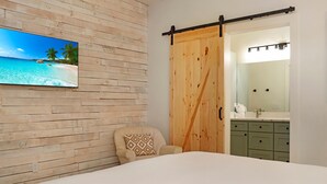 Ein Schlafzimmer mit Holzwand, einem Bett, einem Sessel, einer Schiebetür und einem Badezimmer mit Waschbecken und Spiegel.
