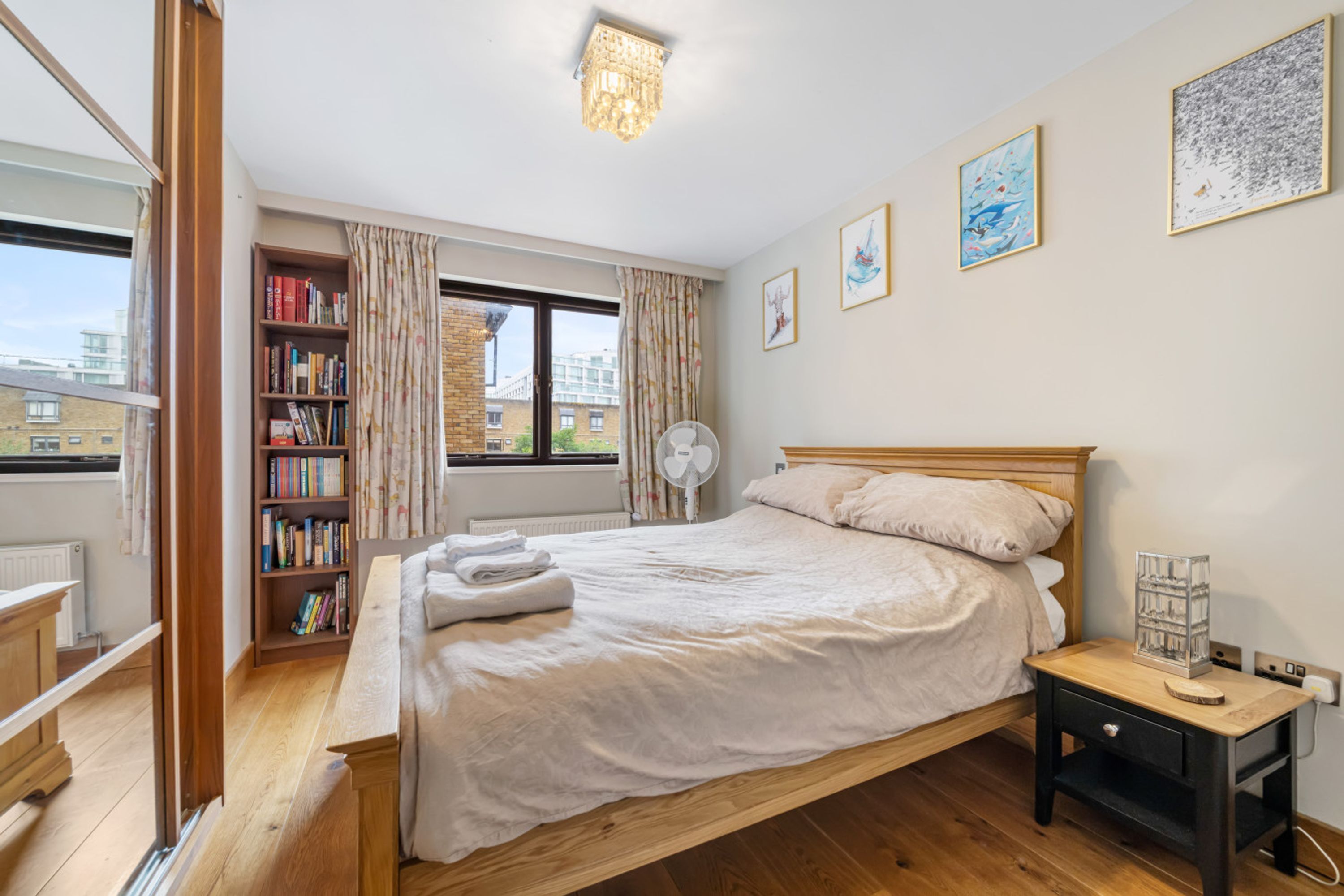 A bedroom with a wooden bed, a bedside table, a bookshelf, a window with curtains, and a view of buildings outside.