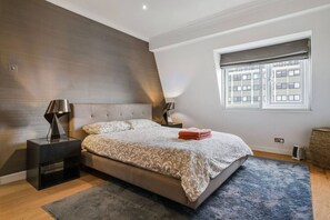A modern bedroom with a large bed, two bedside tables, a window with a view of buildings, and a dark wall. - Spacious Kensington Apartment with City Views (Greater London)