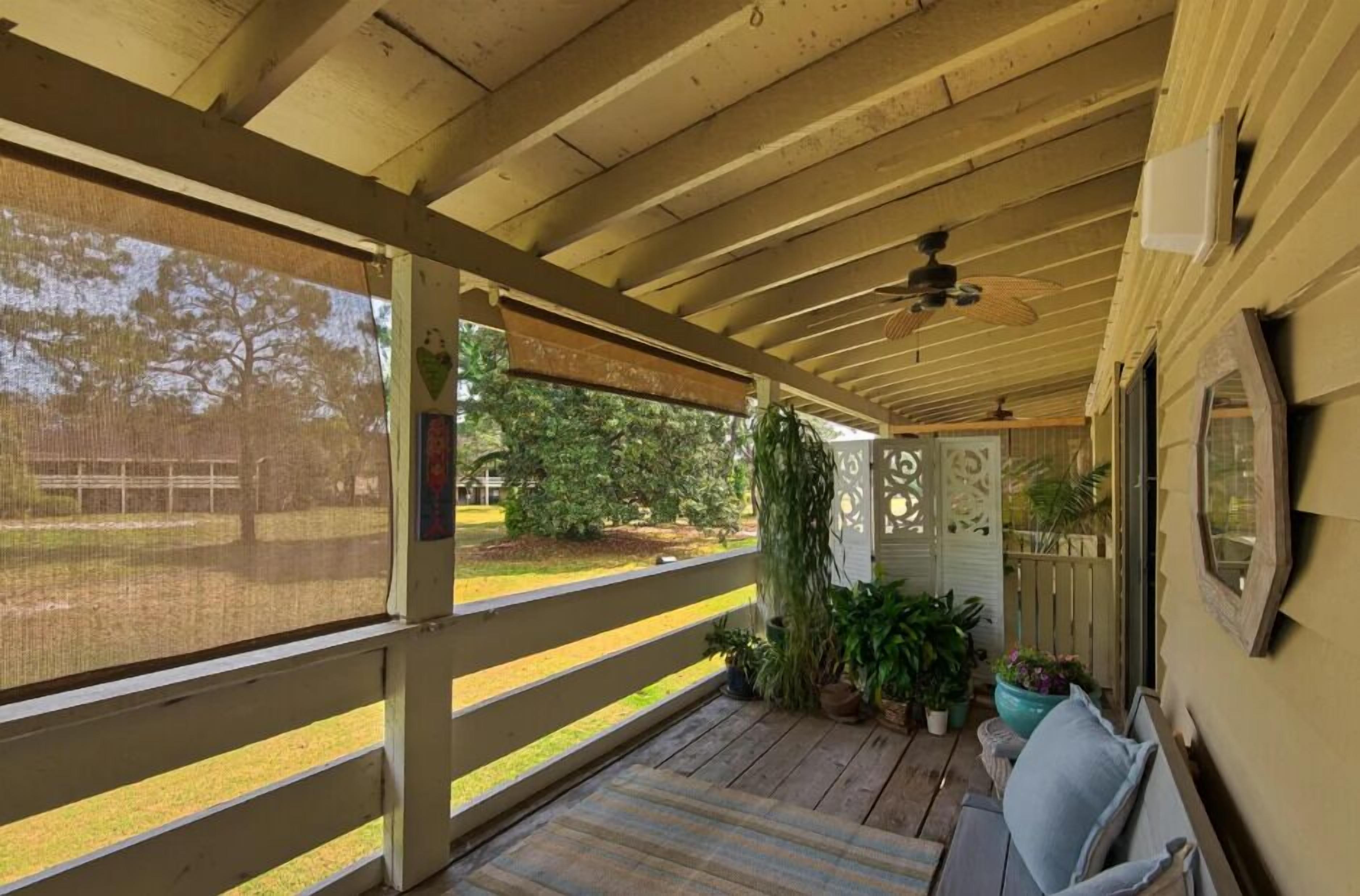 Ein überdachter Balkon mit Holzboden, einem Deckenventilator und Blick auf einen umzäunten Garten mit Bäumen.