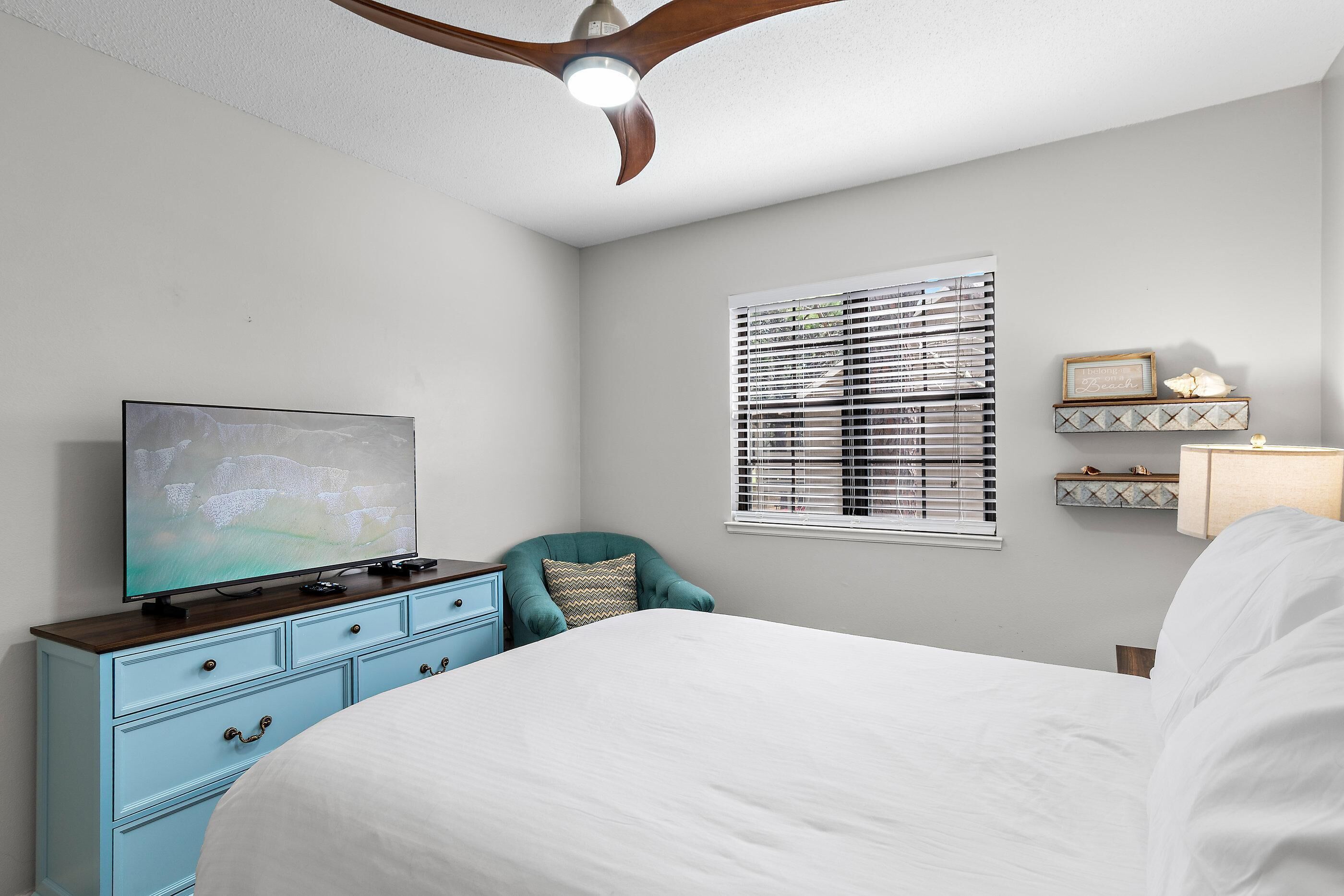 Une chambre à coucher avec un lit, un téléviseur posé sur une commode, un ventilateur de plafond, une fenêtre avec des stores et une table de chevet avec une lampe.