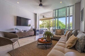 A modern living room with a large flat-screen TV, a sofa, a coffee table, and a large window offering a view of the outdoors. - 3Br-apt | 6 guests | Jacuzzi | Calablanca (Cartagena de Indias)