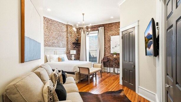 A bedroom with a large bed, a wooden floor, a brick wall, a chandelier, and a TV mounted on the wall.