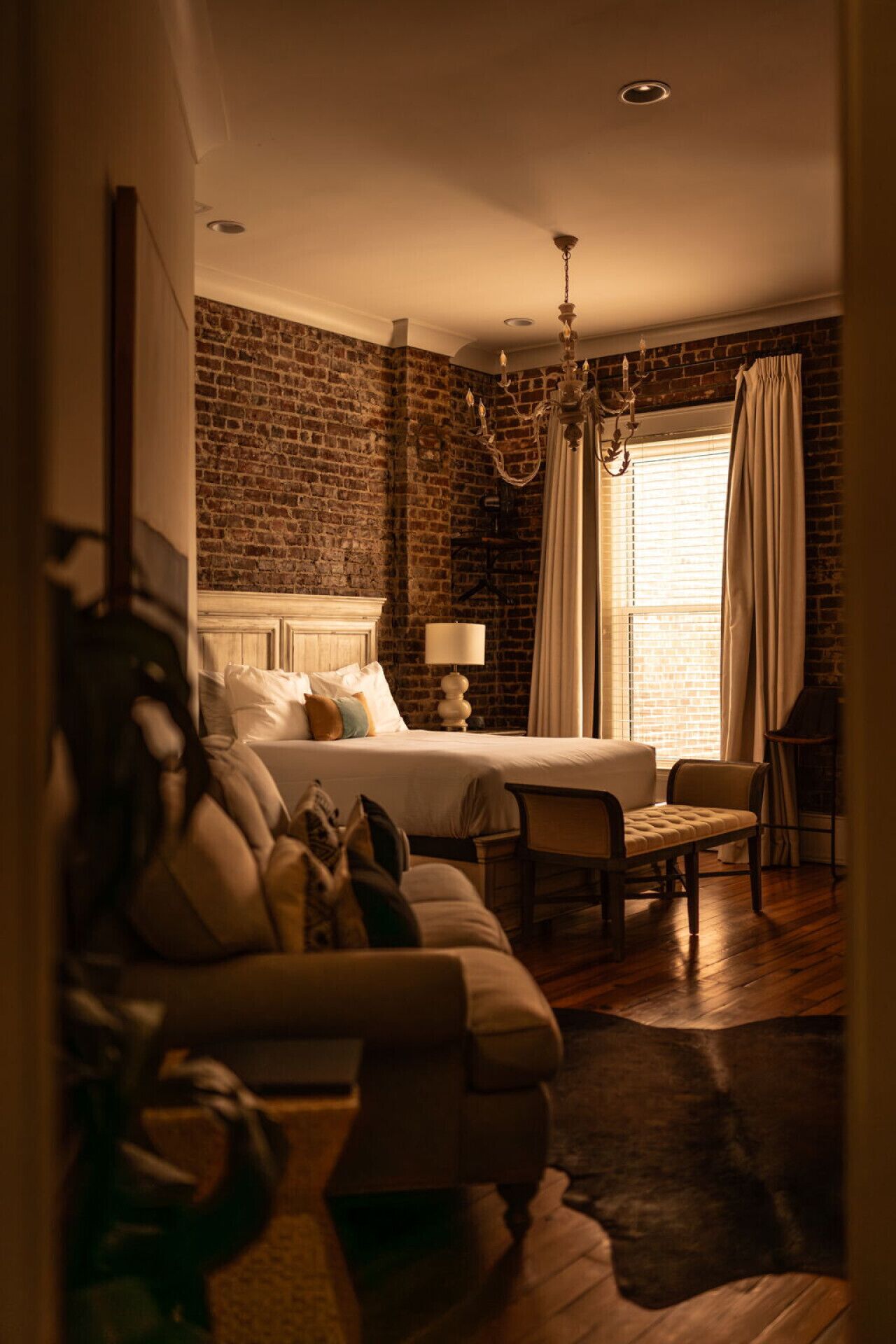 A cozy bedroom with a brick wall, a bed with white linens, a chandelier, and a window with curtains.