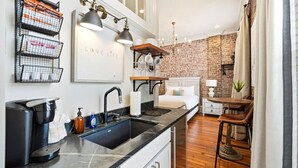 A compact kitchen with a sink, a coffee machine, and a countertop. There is a bed with a white headboard and a small table with chairs. The room has a brick wall and a chandelier.