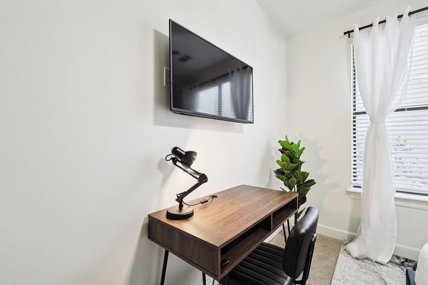 A small, modern room with a wooden desk, a black desk lamp, a television mounted on the wall, a potted plant, and a window with white curtains.