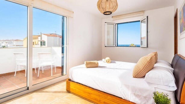 A bedroom with a large bed, a window with a view, a ceiling fan, and a balcony with chairs. - Villa Dreams by Villa Plus (Los Delfines)
