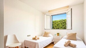 A hotel room with two beds, a chair, a small table, and a window with a view of greenery. - Villa Dreams by Villa Plus (Los Delfines)
