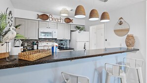 A modern kitchen with white cabinets, a granite countertop, and a bar area with white stools. - Tradewinds Condominium #28- At the Beach! (Miramar Beach)