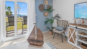 A room with a large sliding glass door leading to an outdoor deck, wicker furniture, a television, and decorative wall hangings. - Tradewinds Condominium #28- At the Beach! (Miramar Beach)