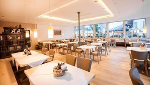 A spacious dining area with multiple tables and chairs, large windows offering a view of the outside, and a modern ceiling design. - Hotel Kendler (St. Gilgen)